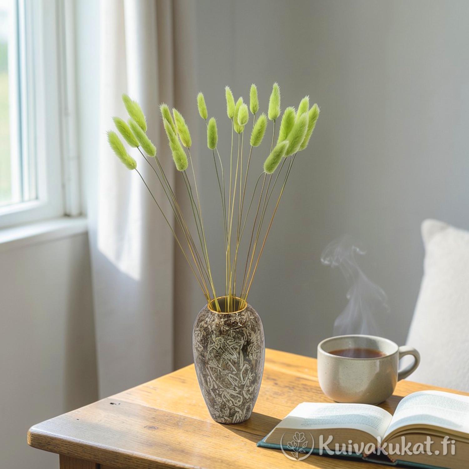 Limenvihreä jänönhäntä (Lagurus ovatus) aseteltuna maljakkoon puisen pöydän päälle, vieressä avattu kirja ja höyryävä kuppi.